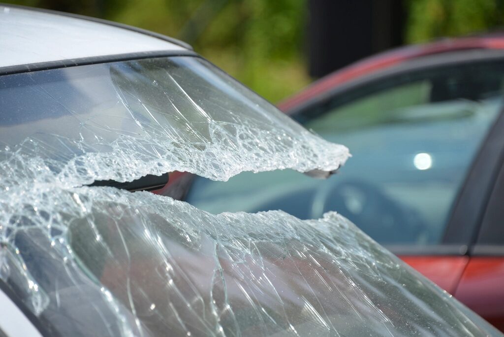 Zersplitterte Windschutzscheibe eines weißen Autos, im Hintergrund ein rotes Fahrzeug